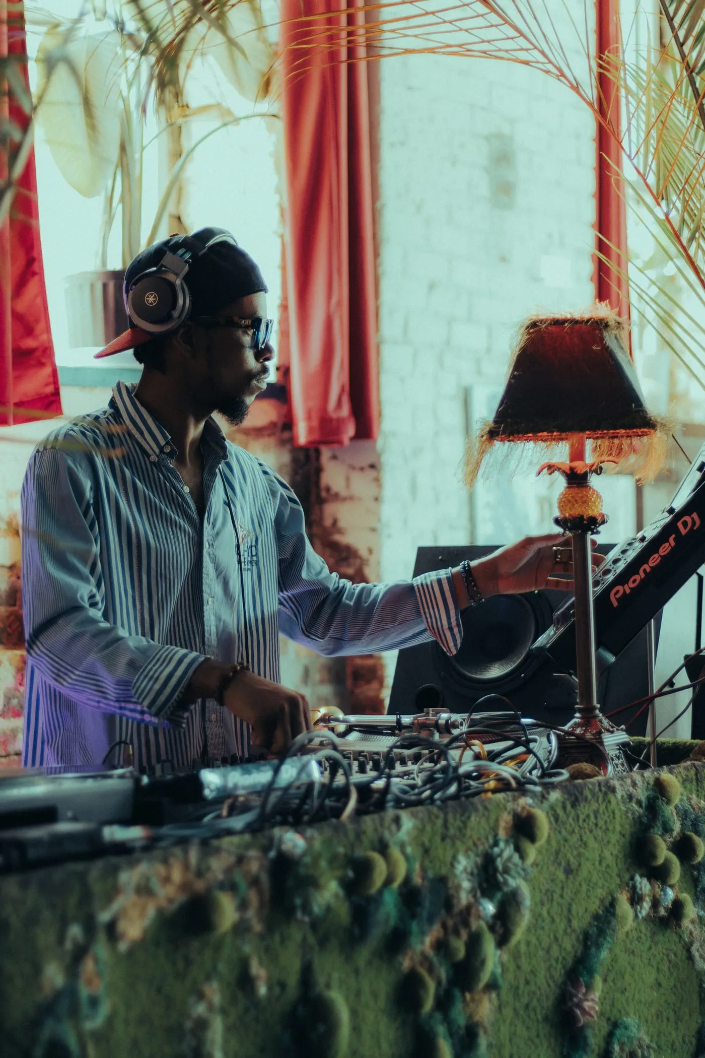 A DJ in headphones working a pair of decks beside a fringed lamp