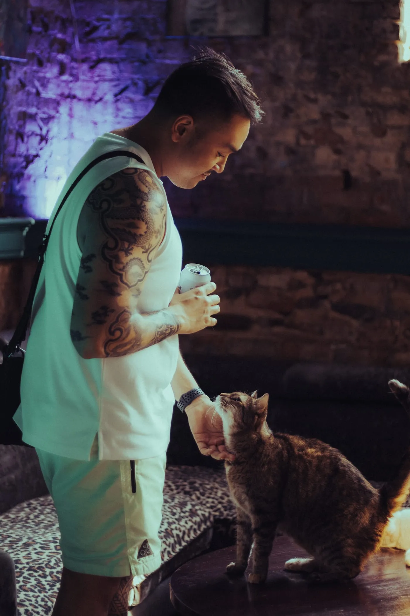 A man holding a drink, crouching to let a tabby cat sniff his hand